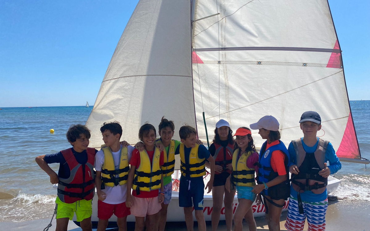 Grupo de niños de 7 a 11 años con chaleco salvavidas junto a un velero en la playa de Oliva
